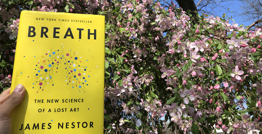 A hand holds a copy of "Breath" By James Nestor in front of a flowering crabapple tree